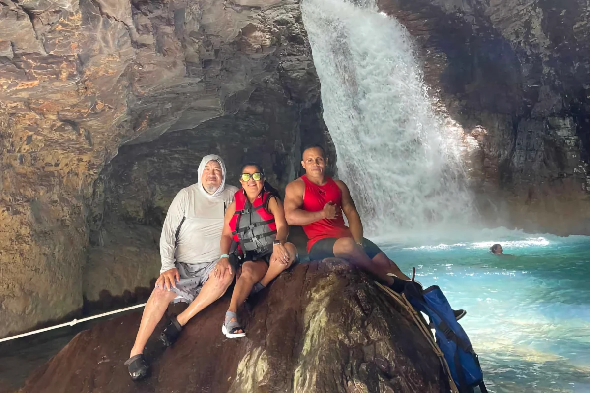 Group of three tour guests sitting on a rock inside the volcanic cave with La Leona Waterfall and turquoise pool in the background, Guanacaste Costa Rica