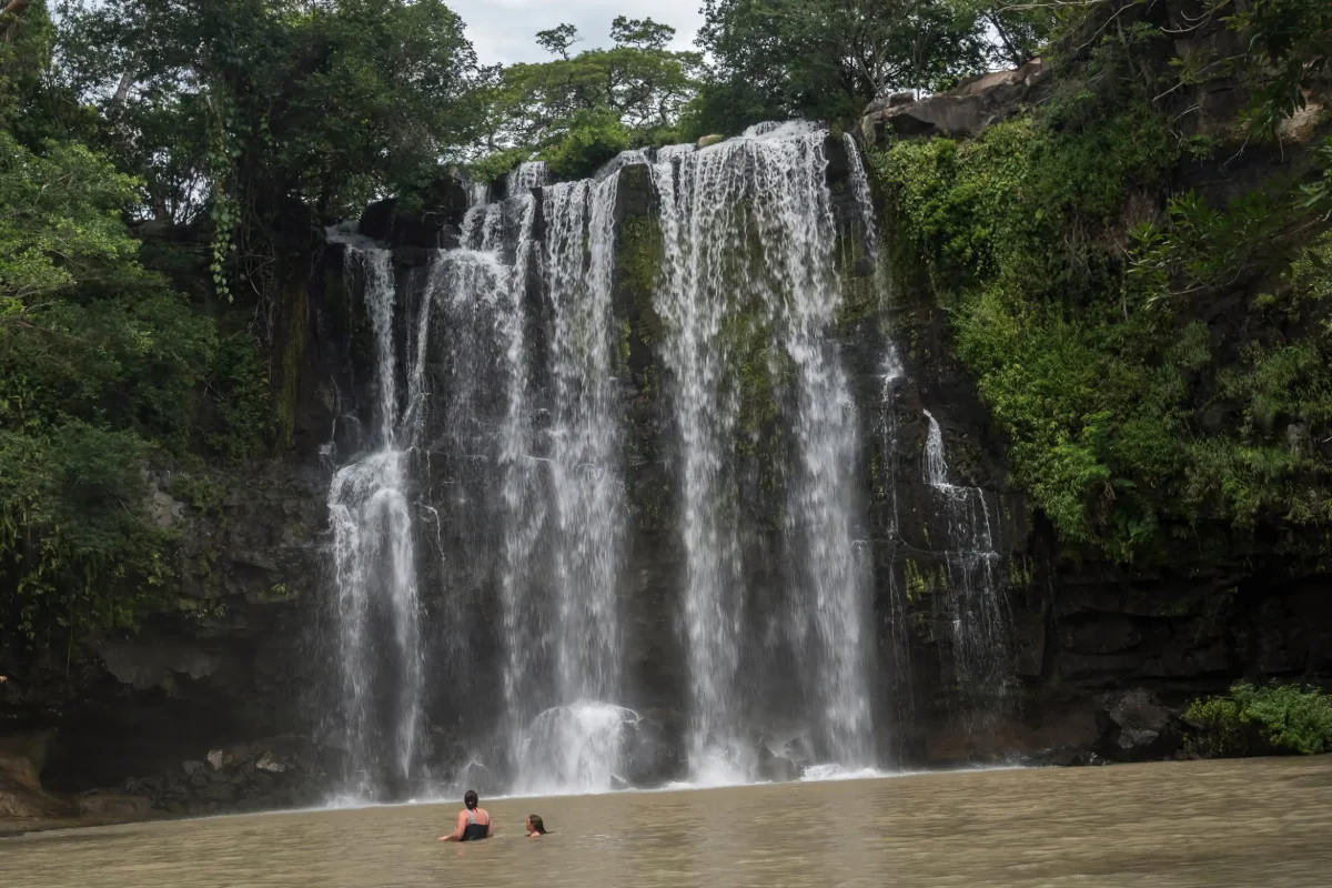 Two guests swimming in the natural pool at the base of Llanos de Cortez waterfall surrounded by tropical jungle in Guanacaste, Costa Rica